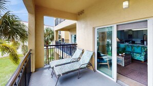 A balcony with two lounge chairs, a sliding glass door leading to a living area with a sofa and a coffee table, and a view of a palm tree and building in the background. - Water view condo with pool hot tub pier private balcony AC and washer dryer (Treasure Island)