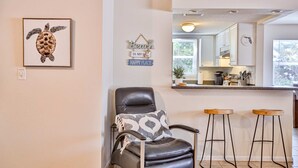 A modern living room with a leather chair, a framed artwork, and a kitchen area visible through an open door. - Quiet Waves 20 Condo with Pool Steps to Dining Shopping and St Pete Beach (St. Pete Beach)