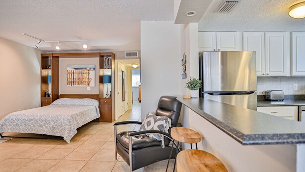 A modern hotel room with a bed, a leather chair, a small bar area, and a kitchenette. - Quiet Waves 20 Condo with Pool Steps to Dining Shopping and St Pete Beach (St. Pete Beach)