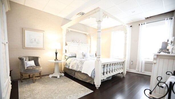 A four-poster canopy bed with a white headboard, a small round table with a lamp, a grey armchair, and a patterned rug.