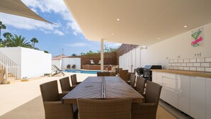 Un espacio exterior para comer con piscina, mobiliario de mimbre y una zona de cocina con parrilla.