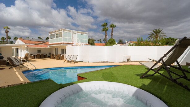 Una piscina con hidromasaje, sillas de descanso y un edificio blanco con tejado rojo.