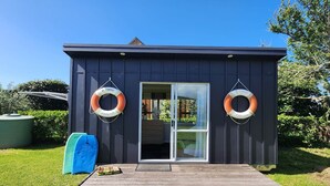 A small, dark-colored building with a wooden deck in front. Two lifebuoys are mounted on the wall. - Seashell Cottage - cosy, newly renovated, central, close to shops and beach (Hahei)