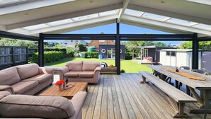 A covered outdoor area with a wooden deck, two sofas, a table with drinks, and a view of a backyard with a house and greenery. - Seashell Cottage - cosy, newly renovated, central, close to shops and beach (Hahei)