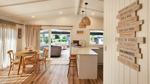 A cozy living room with a dining area, a kitchen, and a large window. - Seashell Cottage - cosy, newly renovated, central, close to shops and beach (Hahei)