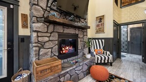 A stone fireplace with a TV above it, a striped armchair, and a sign for wood pallets. - Romantic Cabin | Hot Tub · Gas Grill · Fireplace (Broken Bow)