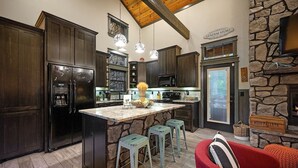 A kitchen with dark wooden cabinets, a central island with stools, a stone fireplace, and a sign reading "General Store." - Romantic Cabin | Hot Tub · Gas Grill · Fireplace (Broken Bow)