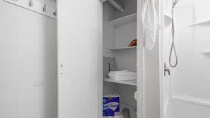 A white bathroom with a shower, a towel rack, and a shelf with folded towels and toilet paper.