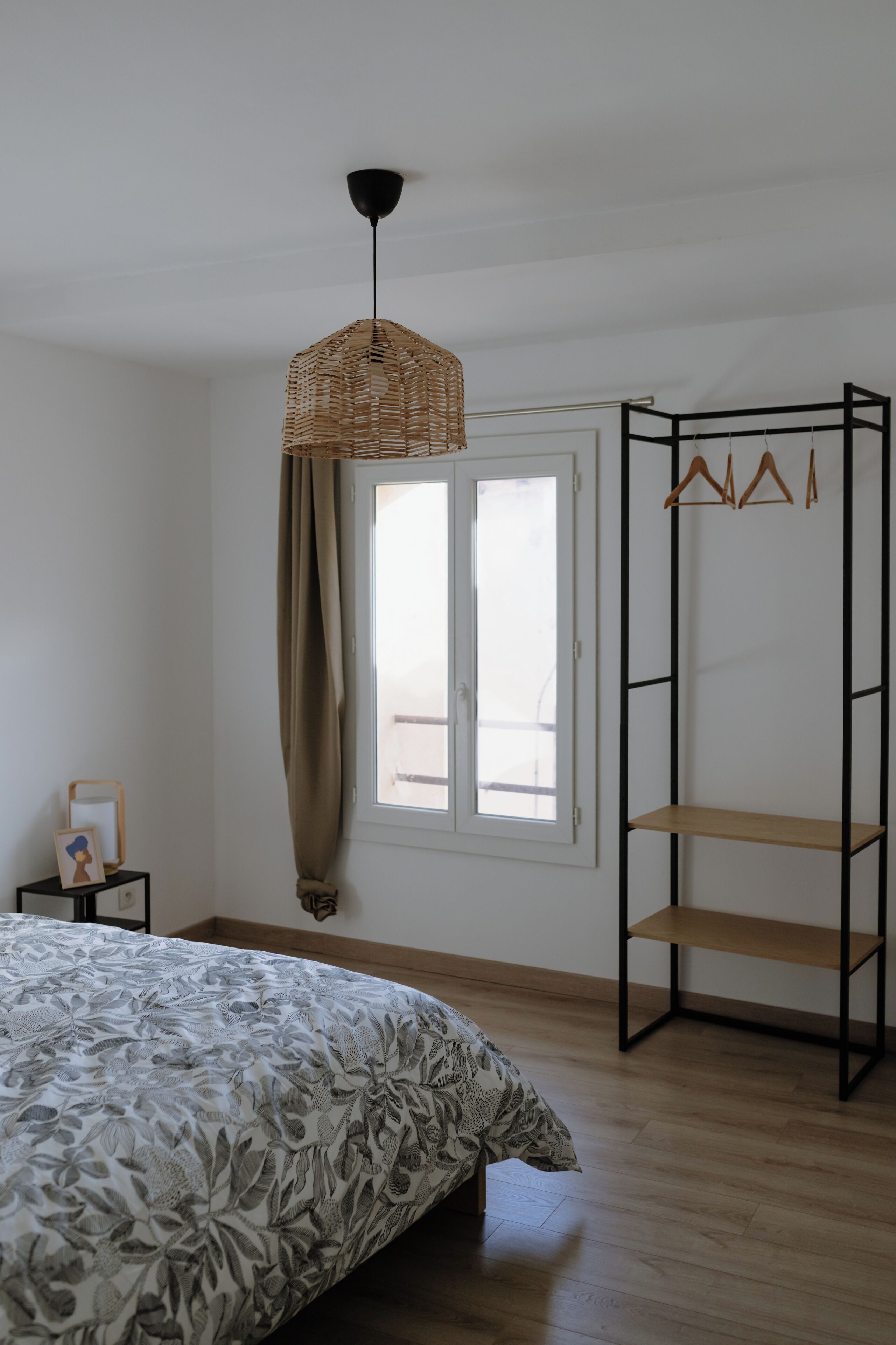 A bedroom with a bed, a hanging light fixture, a window with curtains, a coat rack, and a wooden floor.