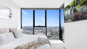 A bedroom with a large window offering a city view, a bed with a blanket, and a painting on the wall. - Comfy 1 Bedroom Unit with River/City View - Q Stay (Broadbeach)