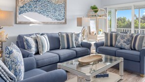 A living room with two blue sofas, a glass coffee table, and a large abstract painting on the wall. - Stunning West End Sanibel Condo – King Suite, Pool, Tennis & Private Beach Acces (Sanibel)