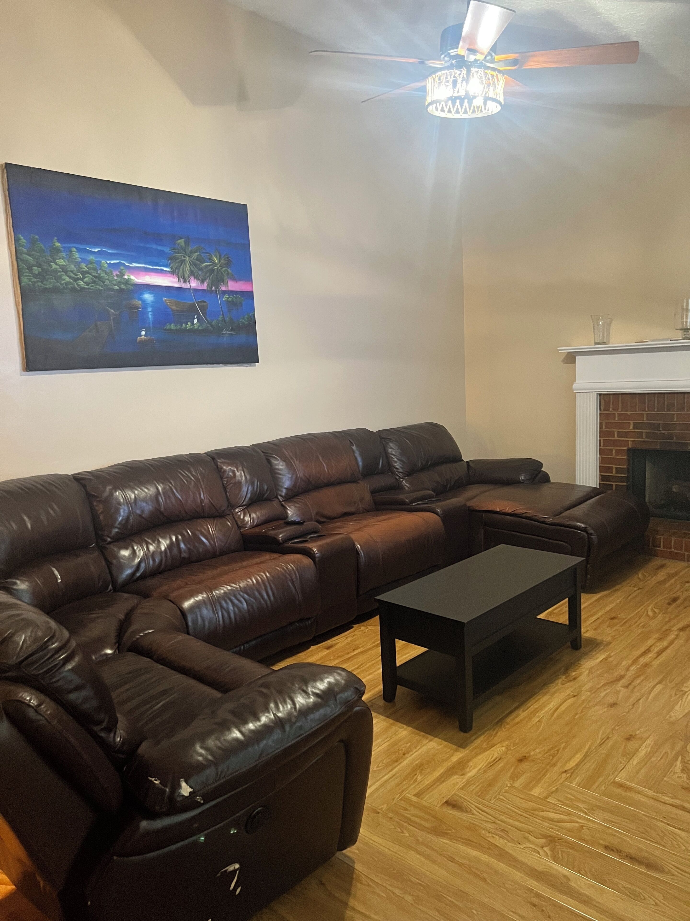 Un salon avec un canapé d’angle en cuir marron, une table basse en bois foncé et un tableau représentant une scène tropicale accroché au mur.