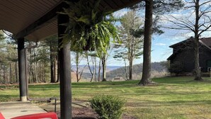 Un espacio al aire libre techado con un banco de madera, un arbusto pequeño y vistas a una cabaña en un paisaje boscoso.