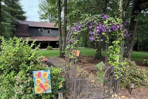 A garden with a metal arbor covered in purple flowers, surrounded by greenery and a house in the background.