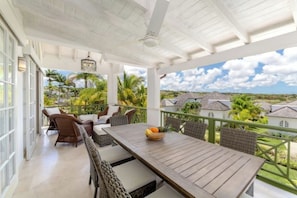 A covered outdoor patio with a wooden table and wicker chairs, overlooking a tropical landscape with palm trees and houses.