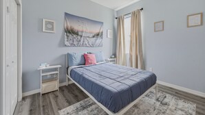 Un dormitorio con cama, mesita de noche, repisa en la pared, ventilador de techo y ventana con cortinas.