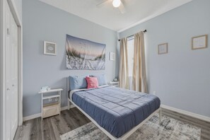 Ein Schlafzimmer mit einem Bett, einem Nachttisch, einer Wandablage, einem Deckenventilator und einem Fenster mit Vorhängen.