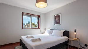A neatly made bed with white linens and folded towels, a bedside table with a lamp, a framed picture on the wall, and a window with a view of buildings. - The Porto Wine Cellars Penthouse (Vila Nova de Gaia)