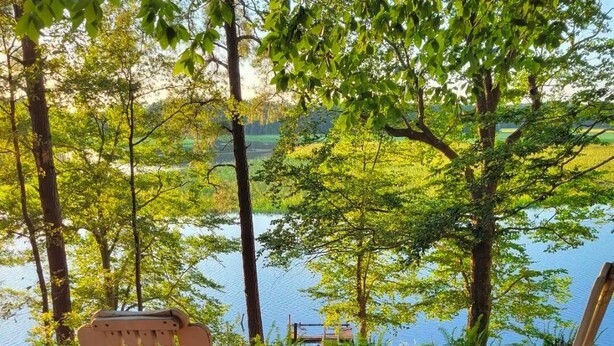 Due poltrone Adirondack con cuscini, un tavolino e un cesto, su un pontile di legno con vista su un lago, alberi e un molo sullo sfondo.