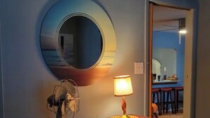 Ett sovrum med en säng, en byrå, en lampa, en rund spegel och utsikt in i ett annat rum.