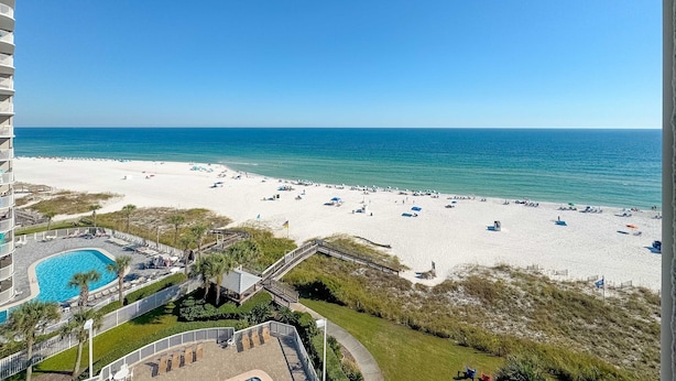 Vista da praia a partir de uma varanda de hotel, com piscina, guarda-sóis e céu claro.