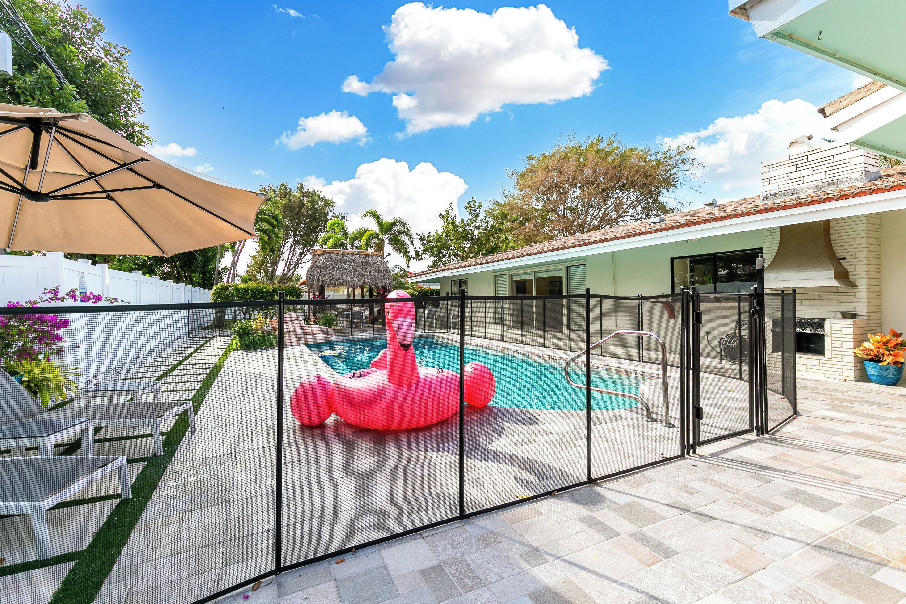 Una piscina con un flotador de flamenco rosa, rodeada por un patio cercado con mobiliario exterior y una estructura con techo de paja.