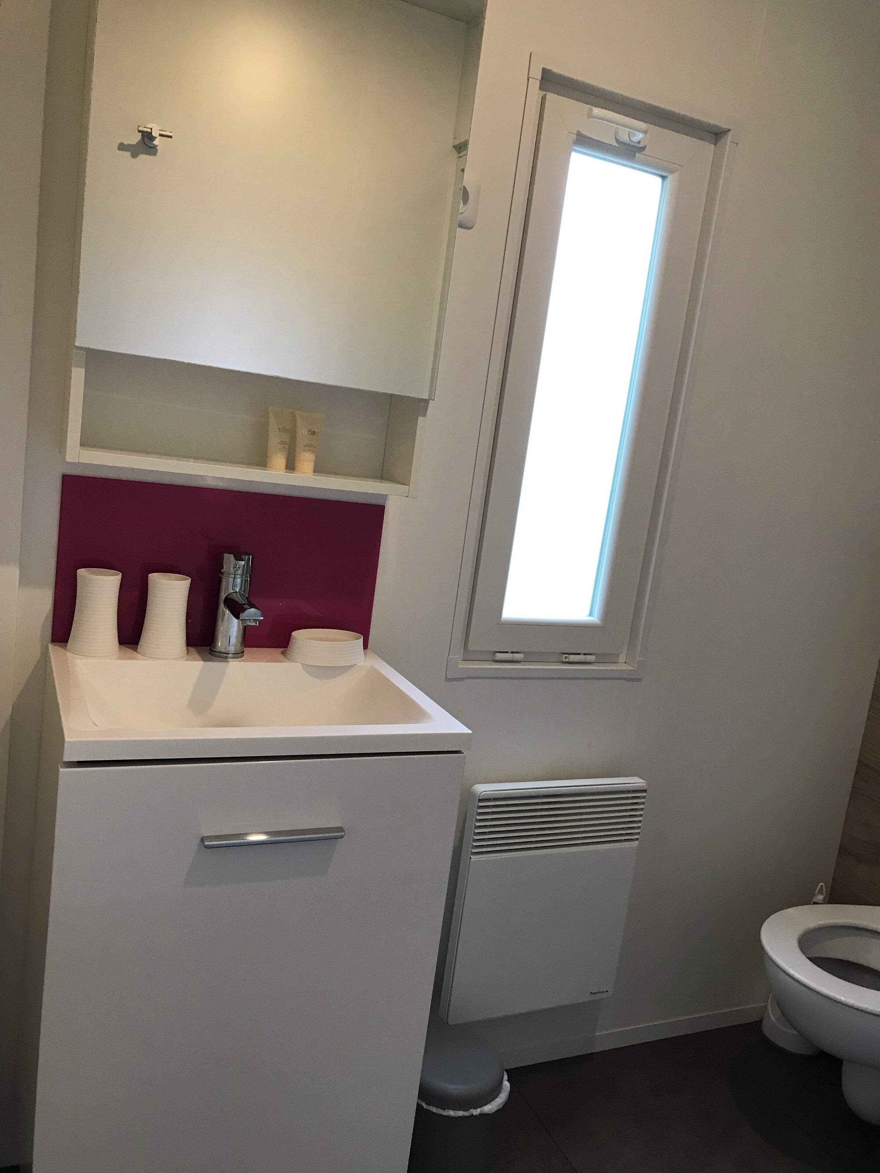 Ein Badezimmer mit einem weißen Waschbecken, einem Spiegel, einem Fenster und einer Toilette.