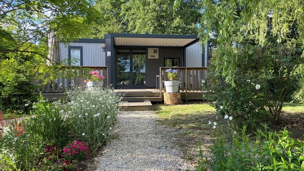 Eine moderne Holzhütte mit einer Terrasse, umgeben von einem Kiesweg und verschiedenen Pflanzen.