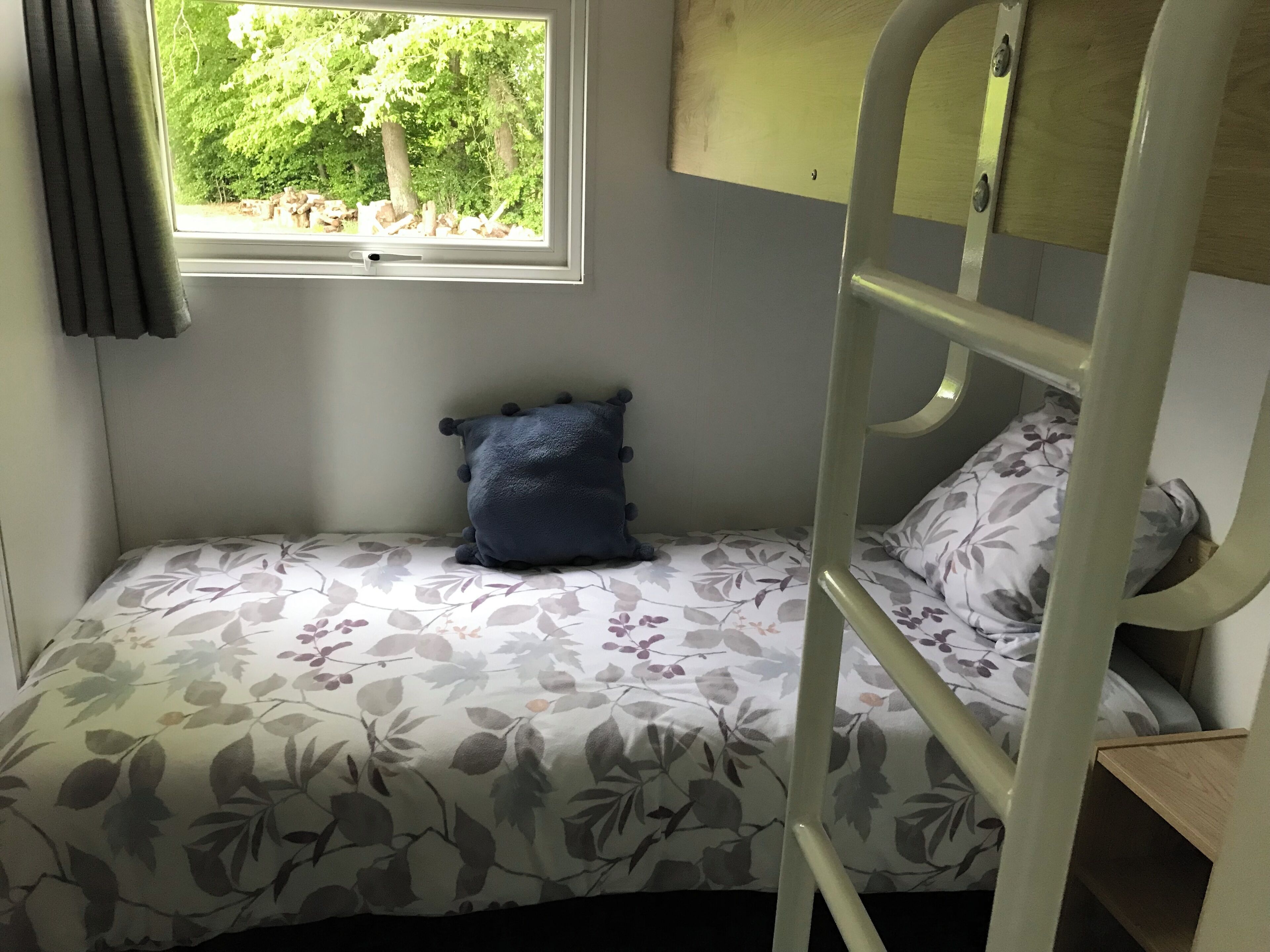 Una cama litera con colcha de estampado floral, un almohón azul oscuro y una ventana con vistas a árboles.