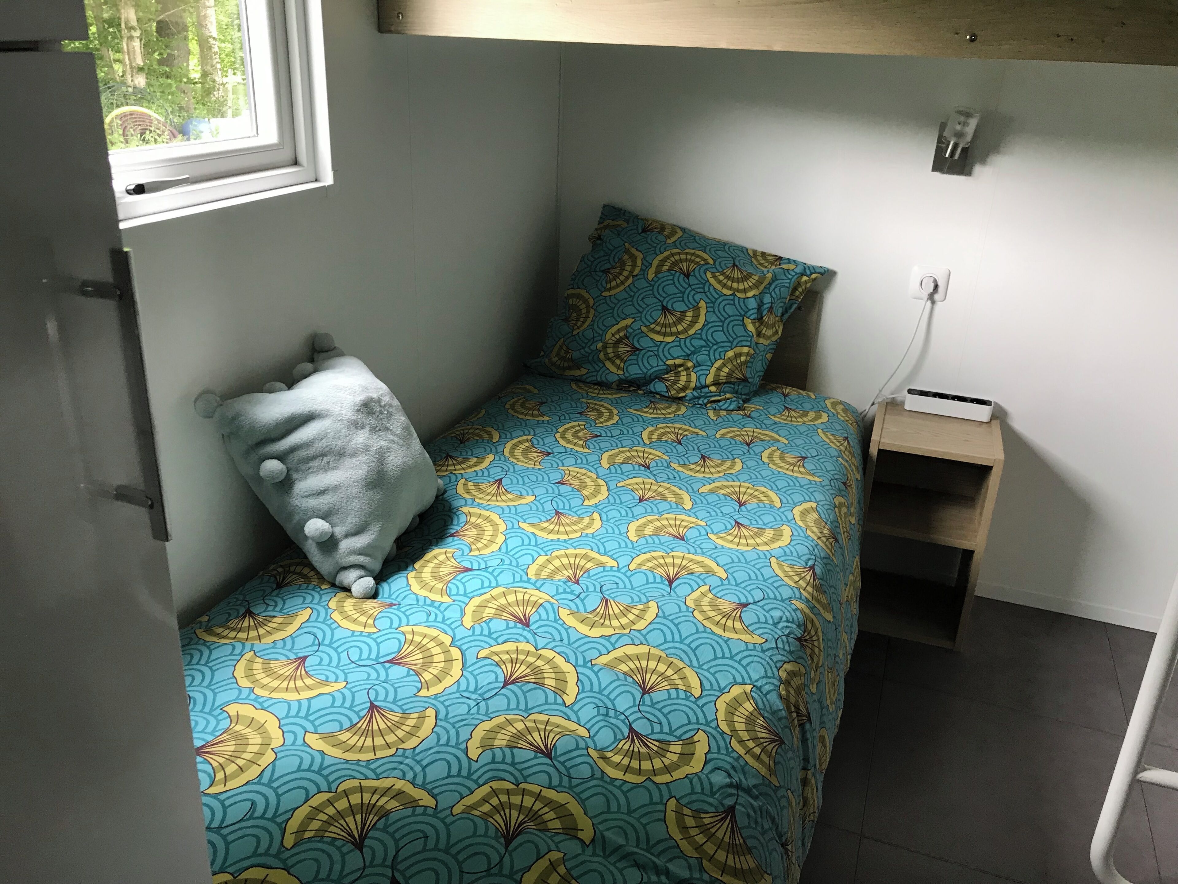 A small, single-bed room with a blue and yellow patterned bedspread, a grey pillow, a bedside table with a power strip, and a window with a view of greenery outside.