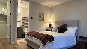 A bedroom with a bed, a bedside table with a lamp, a wall-mounted picture, and a door leading to a bathroom. - New Cozy Fully Furnished Studio Lone Tree (Lone Tree)