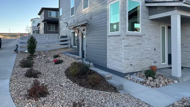 A modern two-story residential building with a grey brick facade, a small front garden with rocks and shrubs, and a white picket fence. - New Cozy Fully Furnished Studio Lone Tree (Lone Tree)