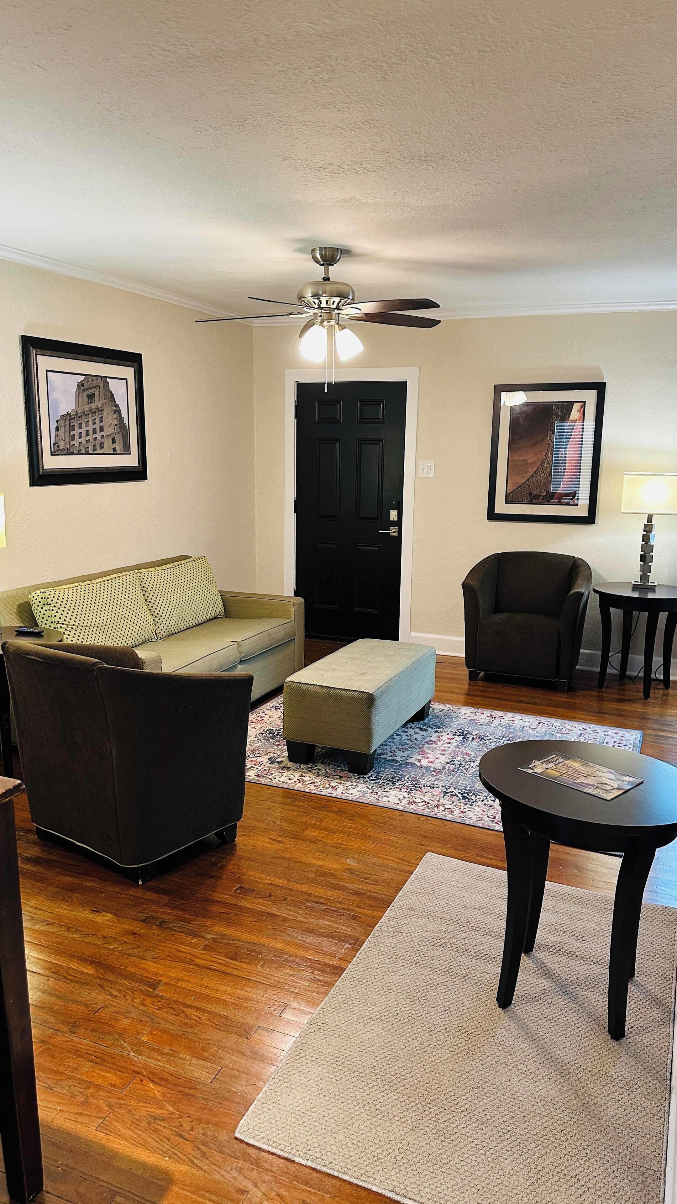 A living room with a sofa, ottoman, armchair, and a small table. There is a ceiling fan, a door, and two framed pictures on the walls.
