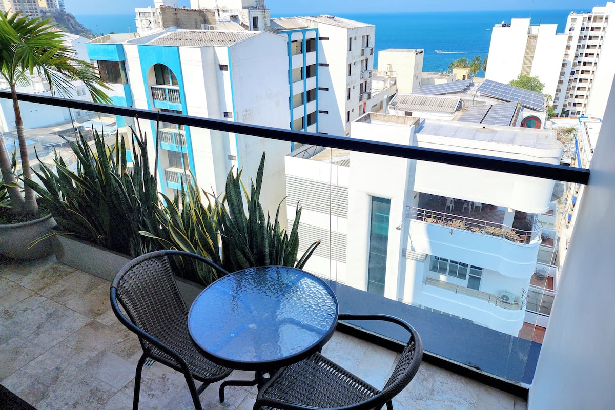 Un balcon avec une table en verre et deux chaises, offrant une vue sur un paysage urbain avec des immeubles et la mer.