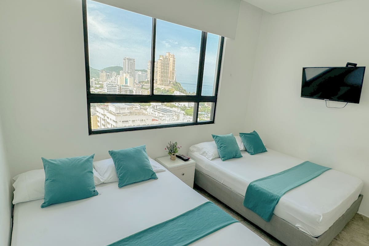 Une chambre d’hôtel avec deux lits, chacun agrémenté de draps et d’oreillers de couleur turquoise, une table de chevet avec une plante et un téléviseur fixé au mur.