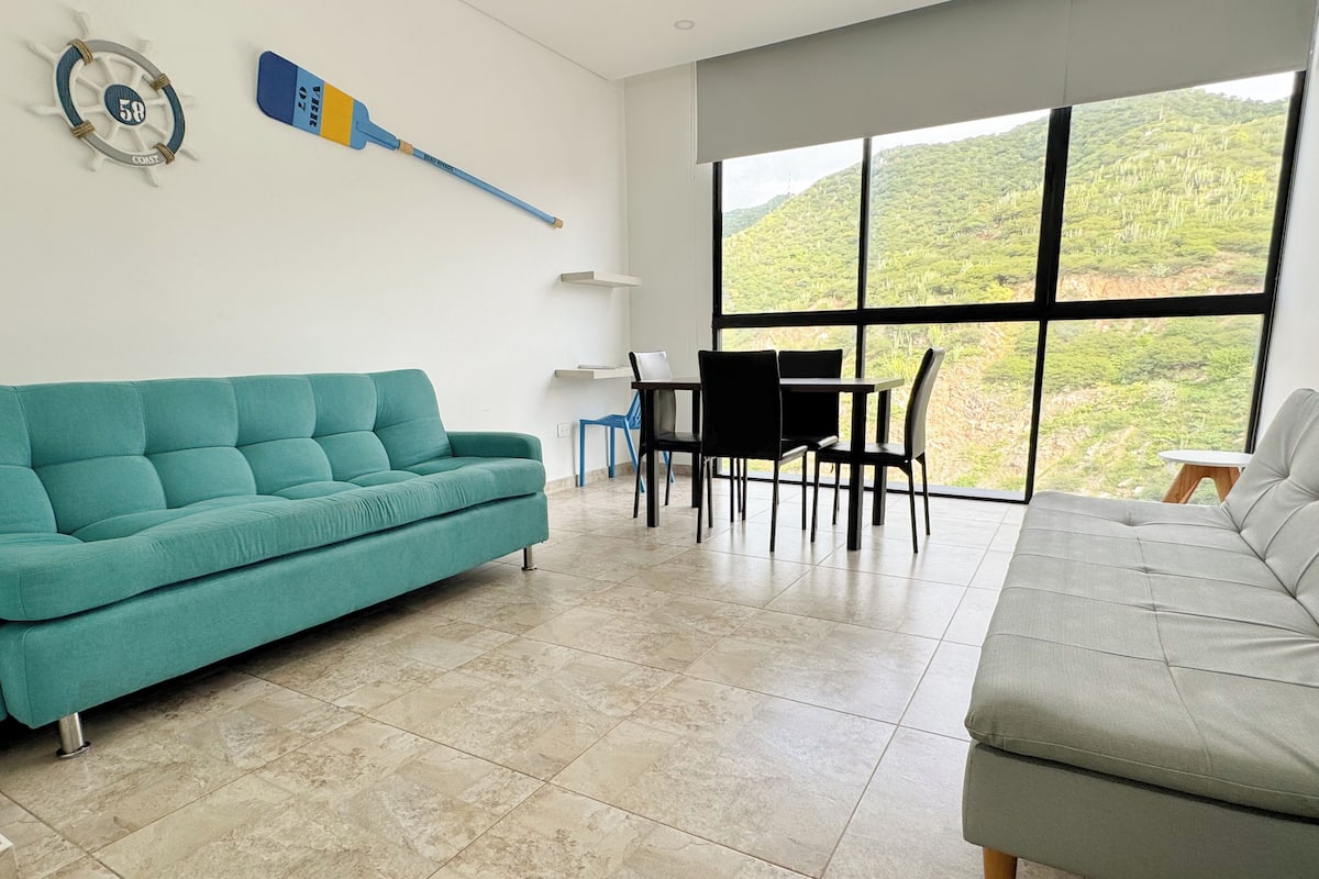 Un salon moderne avec un canapé turquoise, un canapé gris et un coin repas comprenant une table et des chaises. Une grande fenêtre offre une vue sur un paysage verdoyant.