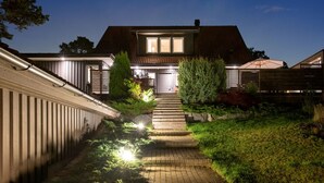 A house with a well-lit entrance, a paved pathway leading to the door, and a small garden area with shrubs and a tree. - Ref. 673271 (Täby)