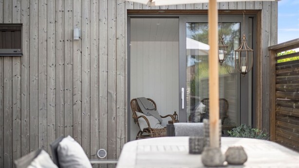 A wooden exterior wall, a glass door, a wicker chair, and a table with a lamp. - Ref. 673271 (Täby)