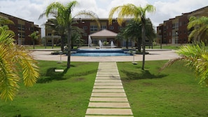 A courtyard with a central fountain, surrounded by palm trees and a pathway leading to a building with multiple floors and balconies.