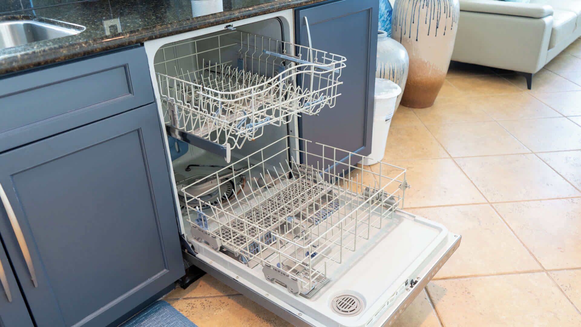 An open dishwasher in a kitchen with grey cabinets and a tiled floor.