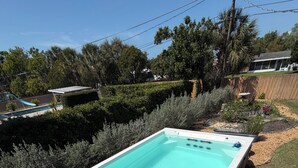 Una bañera de hidromasaje rectangular con borde blanco, rodeada por una terraza de madera, ubicada en un patio trasero con piscina y follaje al fondo.