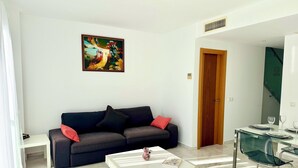 A modern living room with a dark grey sofa, a white coffee table, and a glass dining table with chairs. A colorful abstract painting hangs on the wall, and a wooden door is visible in the background. - Beautiful triplex with sea views in Mijas Costa Calalucia! (Mijas-Costa)
