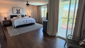 Ein Schlafzimmer mit Bett, Deckenventilator, Nachttisch mit Lampe, Fenster mit Vorhängen und Blick ins Freie.