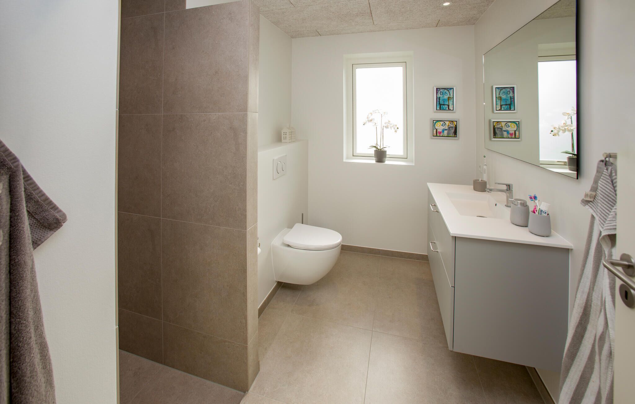 A modern bathroom with a toilet, sink, and mirror. There is a window with a plant and artwork on the wall.