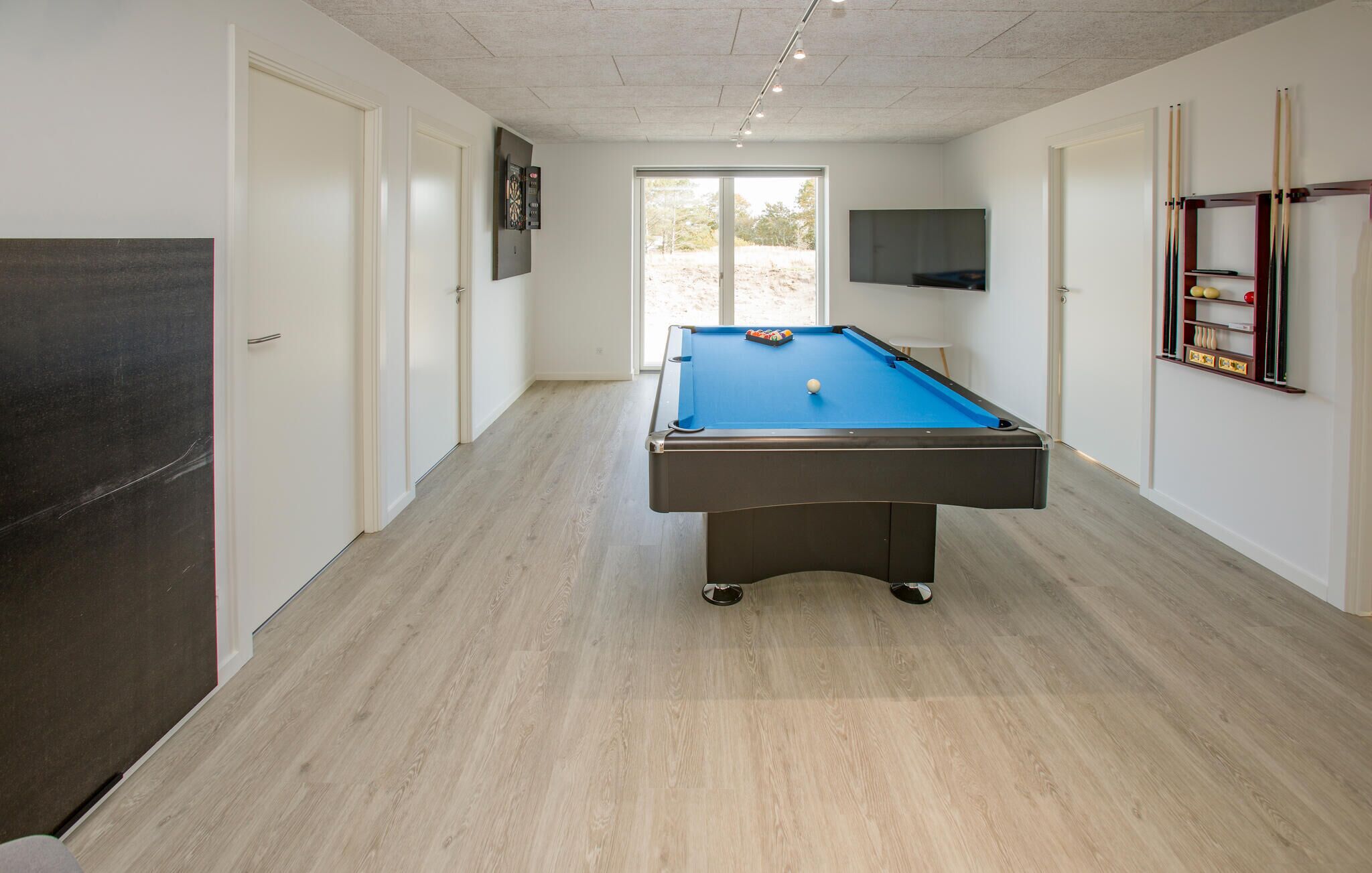 A pool table in a room with a wooden floor, a door on the left, a TV on the wall, and a shelf with pool cues on the right.