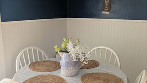 Una mesa redonda de comedor con mantel blanco, rodeada de sillas blancas, un florero con flores y individuales tejidos.