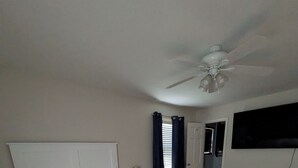 Un dormitorio con una cama grande, una mesita de noche, un ventilador de techo, una ventana con cortinas y una puerta que da a otra habitación.