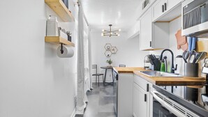 A narrow kitchen with white cabinets, a wooden countertop, and a built-in oven. There is a small dining area with a table and chairs, and a ceiling light fixture.