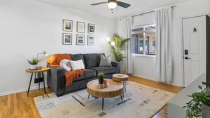 A modern living room with a grey sofa, two round wooden coffee tables, a ceiling fan, and a large window with curtains.