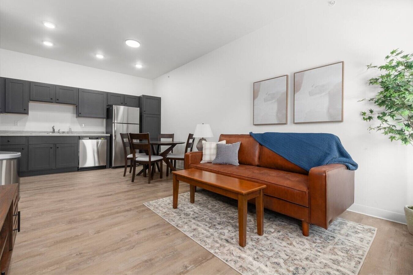 A modern kitchen with grey cabinets and stainless steel appliances, a dining area with wooden chairs, and a brown leather sofa with a blue throw blanket.